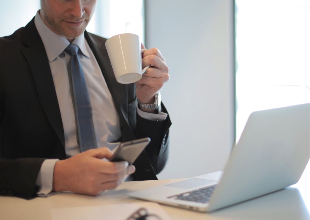 imprenditore guarda il telefono e il computer davanti mentre beve il caffè in ufficio
